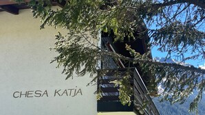 A building with the name 'CHESA KATJA' on its side, a door marked with the number '6', and a view of snow-capped mountains in the background. - 1505 Chesa Katja by Interhome (Valbella)
