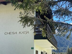A building with the name 'CHESA KATJA' on its side, a door marked with the number '6', and a view of snow-capped mountains in the background. - 1505 Chesa Katja by Interhome (Valbella)