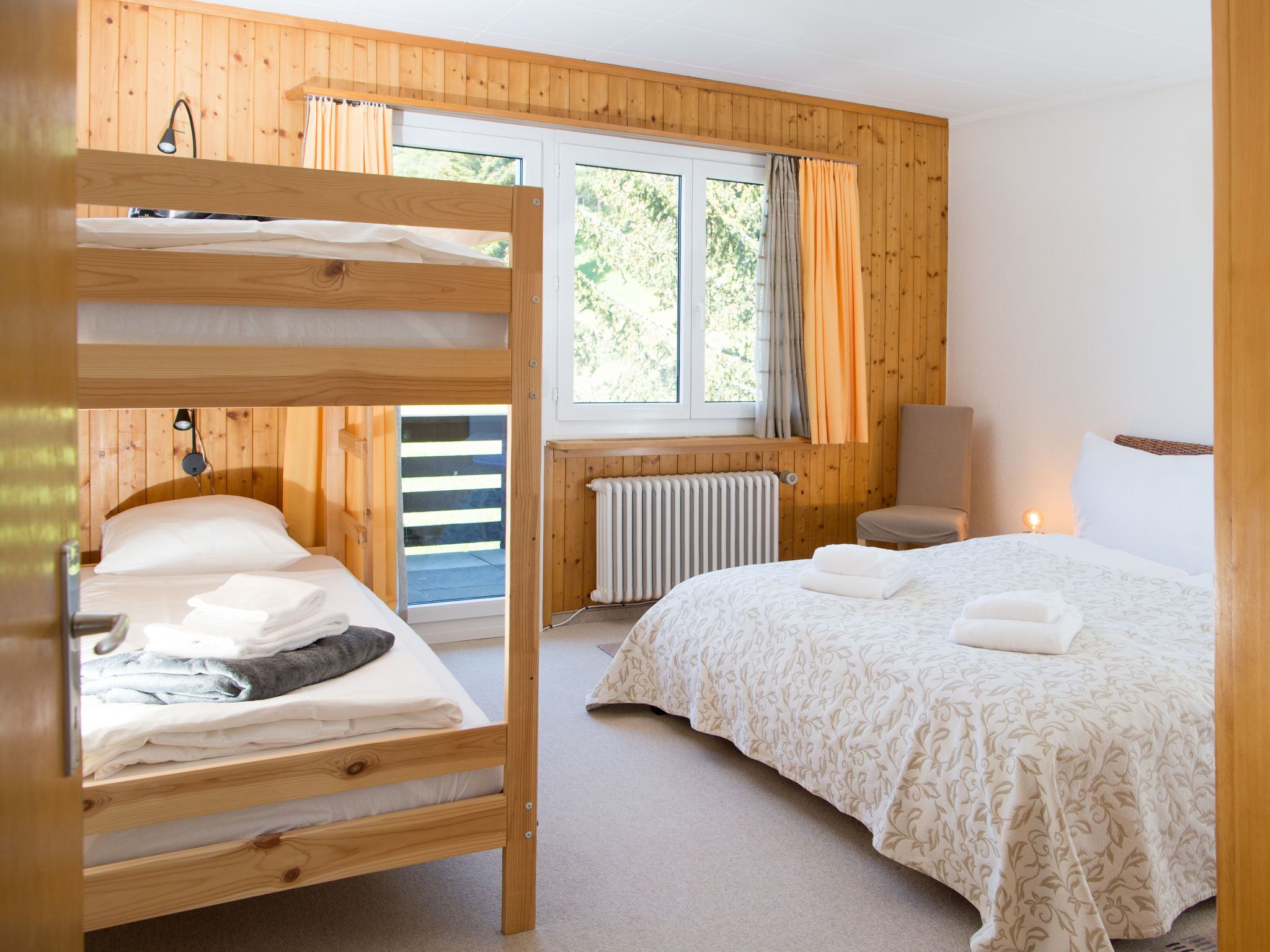 Una habitación con una litera, una cama individual, una silla, un radiador y una ventana con cortinas.