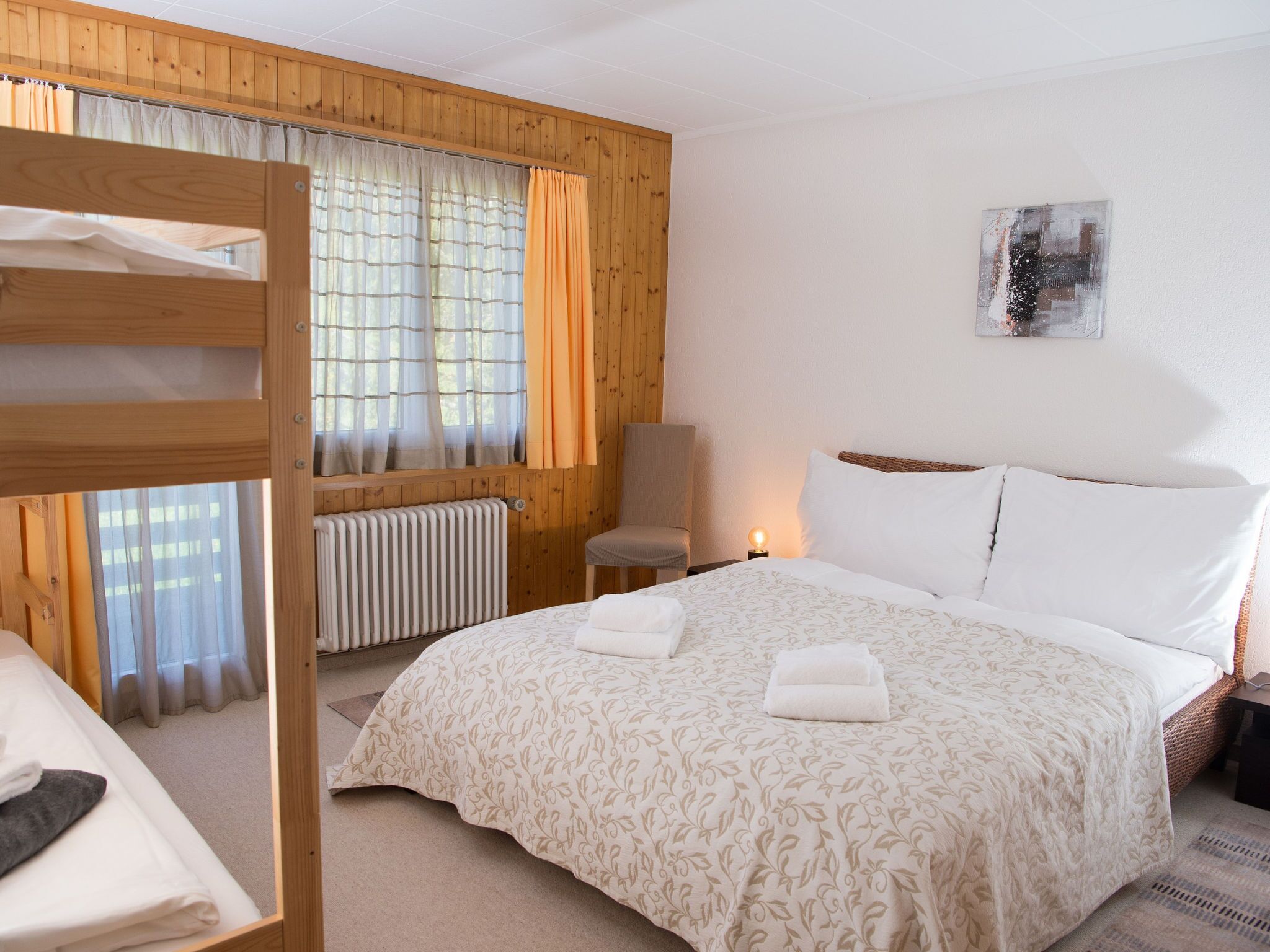 A hotel room with a bunk bed, a single bed with a patterned bedspread, a chair, a bedside table with a lamp, and a radiator.