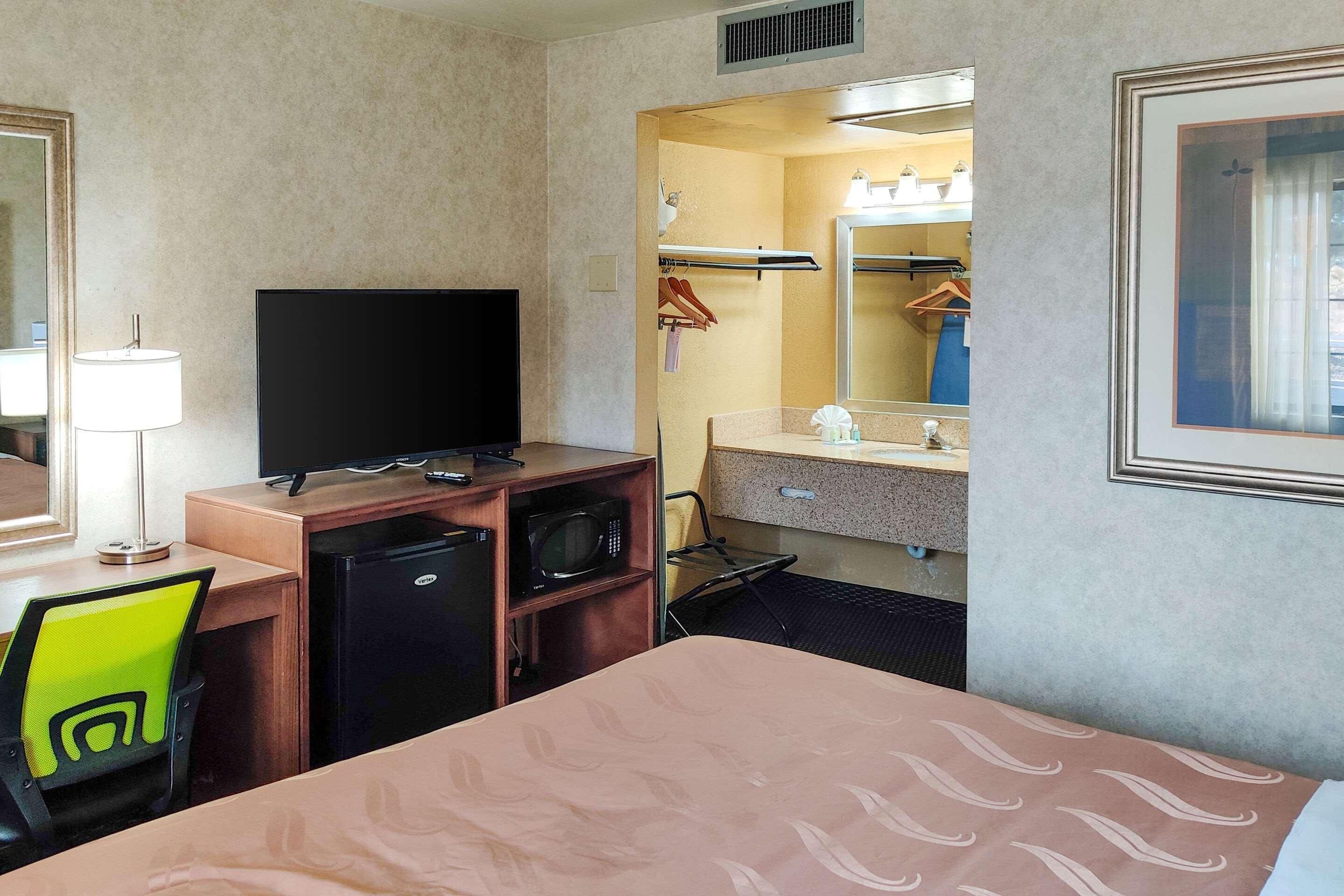 A hotel room with a bed, a desk with a television, a microwave, a small refrigerator, a chair, a mirror, and a bathroom with a sink and towel rack.