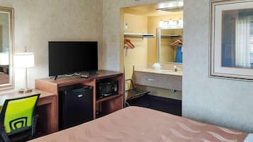 Quarto de hotel com cama, escrivaninha com televisão, micro-ondas, mini-geladeira, cadeira, espelho e banheiro com pia e porta-toalhas.