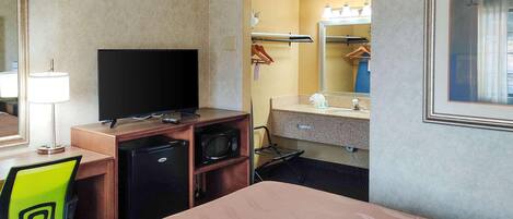 Une chambre d’hôtel comprenant un lit, un bureau avec une télévision, un four à micro-ondes, un petit réfrigérateur, une chaise, un miroir et une salle de bain avec un lavabo et un porte-serviettes.