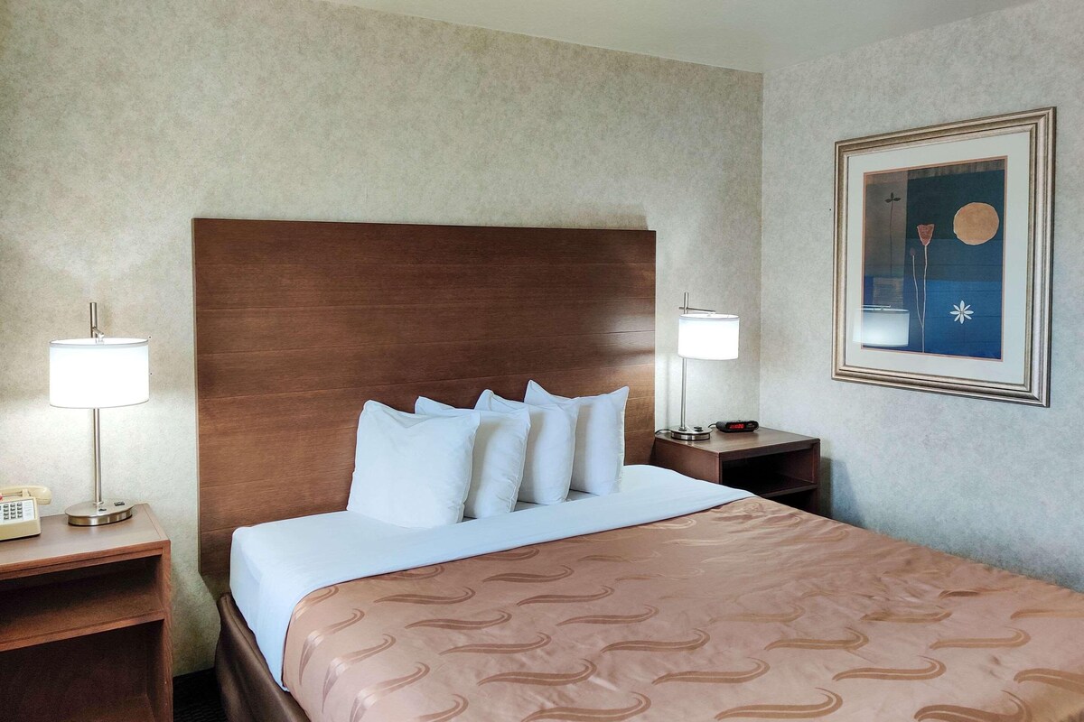 A hotel room with a large bed, two bedside tables, a nightstand, a wall-mounted clock, and a framed picture.