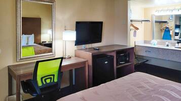 Ein Hotelzimmer mit Bett, Schreibtisch, Fernseher, Kühlschrank und einem Badezimmer mit Waschbecken und Spiegel.