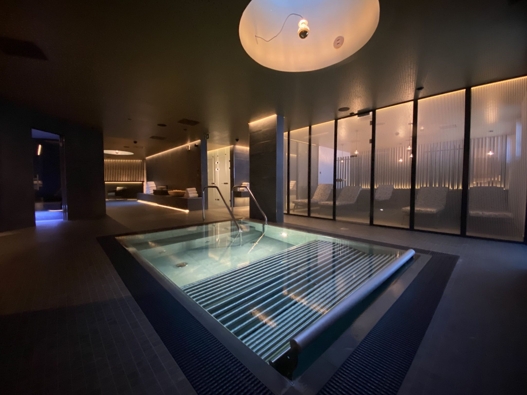 Ein moderner, überdachter Swimmingpool mit Glasbarriere, einem Empfangsbereich und einem großen Fenster mit vertikalen Jalousien.