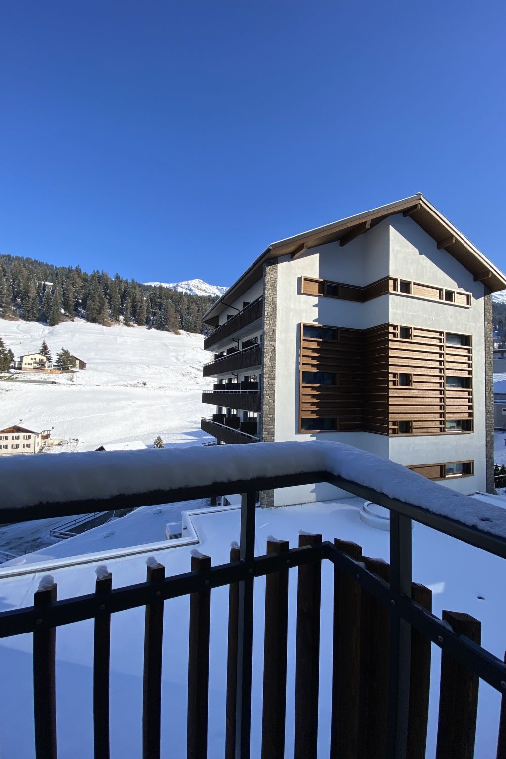 En moderne bygning med balkon, snedækket jord og et bjerglandskab i baggrunden.