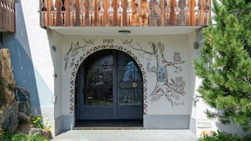Une entrée d’immeuble avec une porte décorative, un balcon doté d’une balustrade en bois et un arbre sur la droite.
