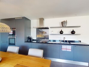 A modern kitchen with grey cabinets, a built-in oven, a stainless steel range hood, and a dining area with a wooden table and chairs. - 1901 Sot by Interhome (Lantsch)