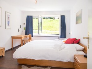 Een slaapkamer met een houten bed, een bureau en een stoel. Er is een raam met gordijnen en een schilderij aan de muur.