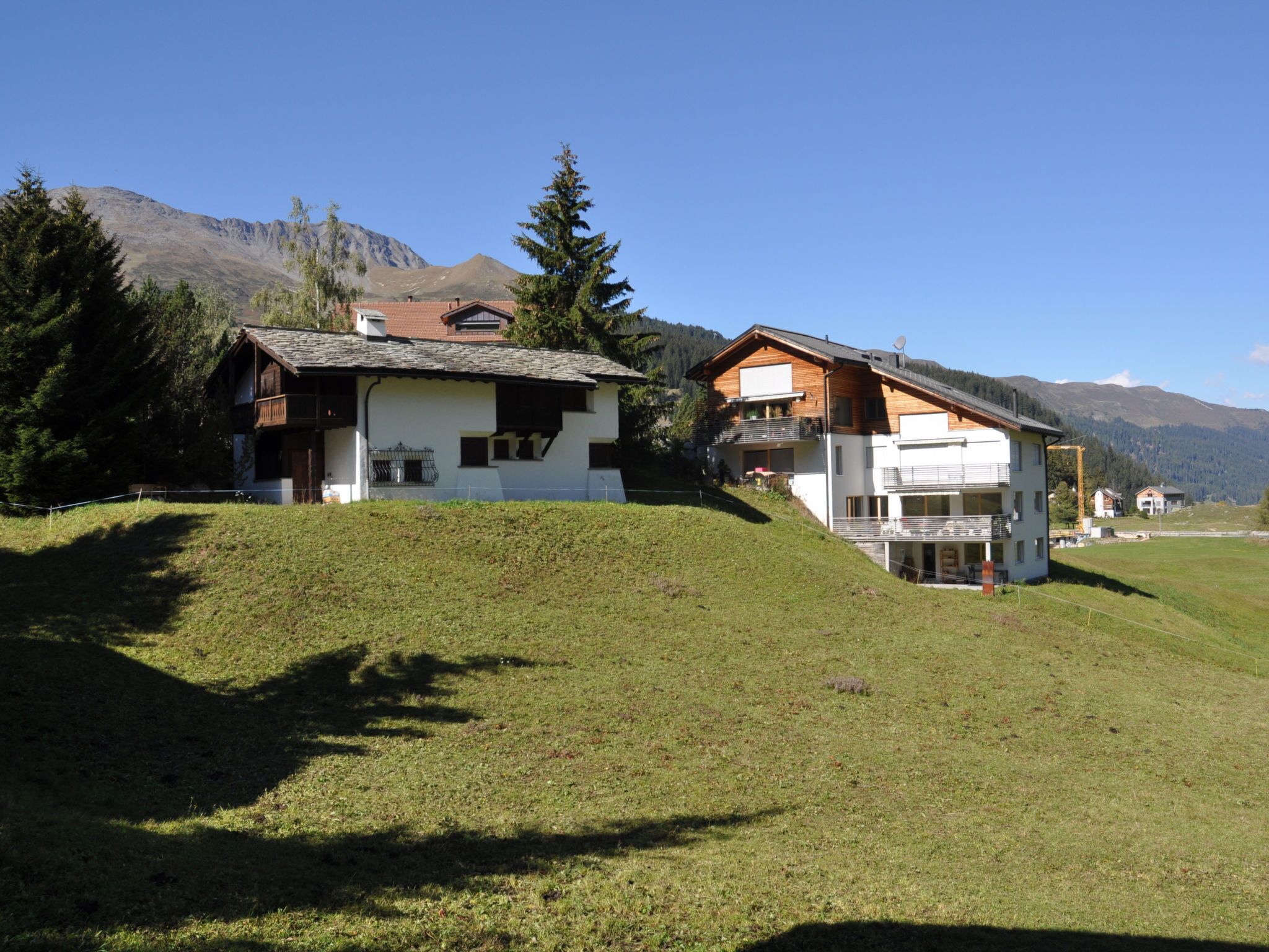 A hillside property with two main buildings, one with a sloped roof and the other with a flat roof, surrounded by trees and mountains.
