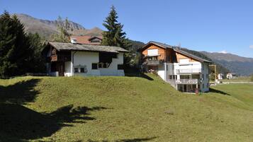 A hillside property with two main buildings, one with a sloped roof and the other with a flat roof, surrounded by trees and mountains.