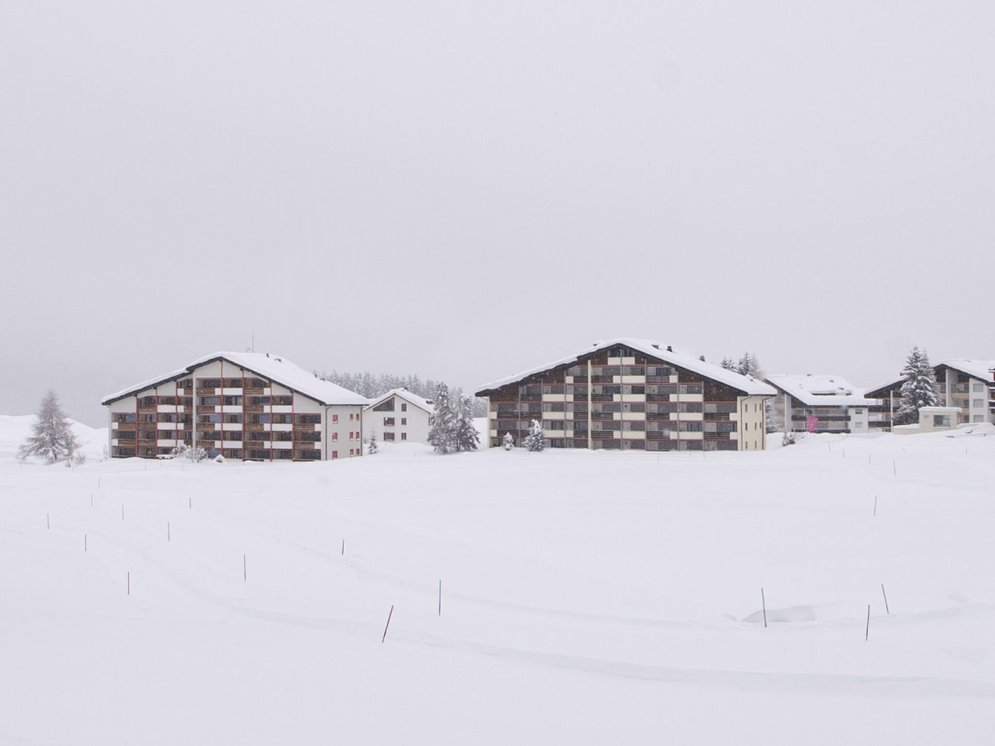 Schneebedeckte Landschaft mit zwei mehrstöckigen Gebäuden, vermutlich ein Hotel oder Resort, mit Balkonen und einer flachen Dachkonstruktion.
