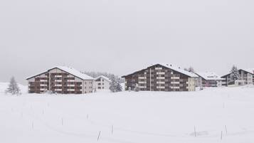 Schneebedeckte Landschaft mit zwei mehrstöckigen Gebäuden, vermutlich ein Hotel oder Resort, mit Balkonen und einer flachen Dachkonstruktion.
