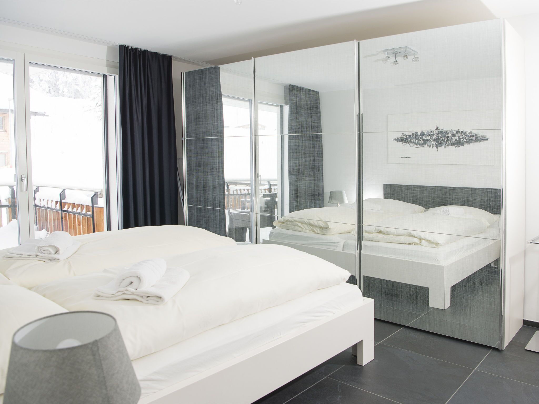 A modern hotel room with a large bed, a separate sleeping area, a balcony with a view, and a cityscape artwork on the wall.
