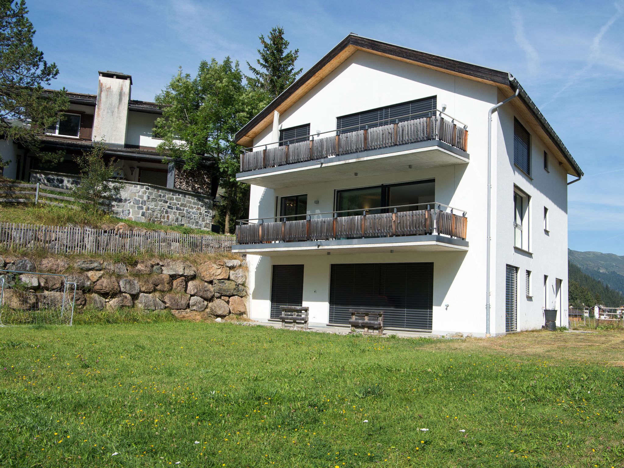 A modern multi-story building with a white facade, balconies, and a stone retaining wall.