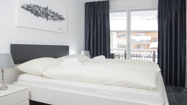 A modern hotel room with a large bed, a bedside table with a lamp, a view of the city through the window, and a painting on the wall. - 1401 Heimberg by Interhome (Parpan)