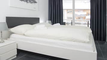 Quarto de hotel moderno com uma cama grande, uma mesa de cabeceira com abajur, vista da cidade pela janela e um quadro na parede.