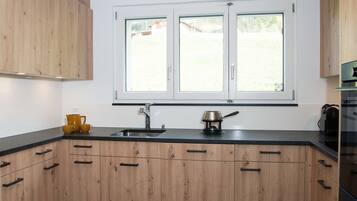 Une cuisine avec des meubles en bois, un plan de travail noir, un évier à deux bacs et une fenêtre donnant sur une maison et des arbres.