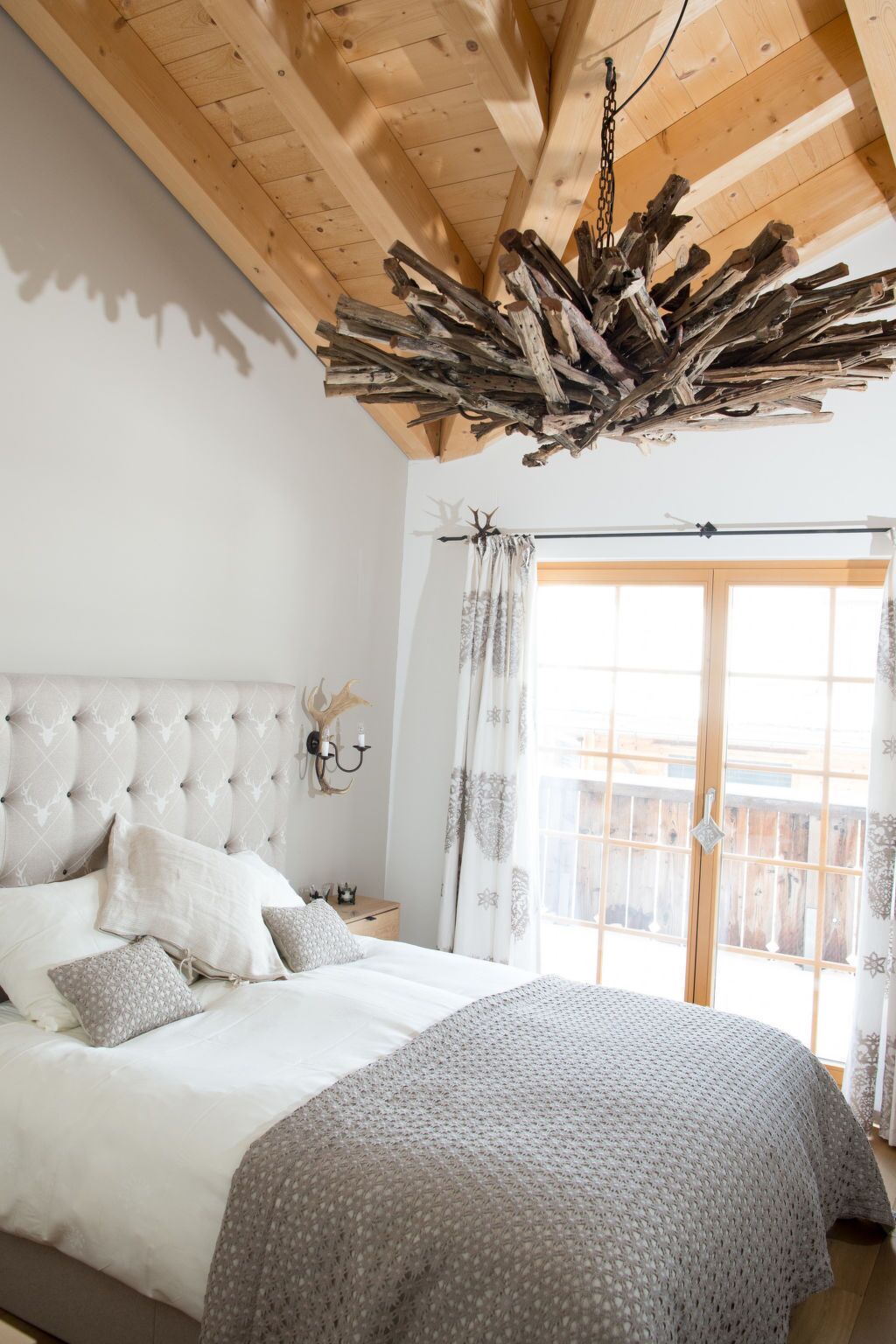 Ein Schlafzimmer mit einer gesteppten Kopfteil, einem Bett mit weißer und gemusterter Bettwäsche, einer Holzbalkendecke und einem Fenster mit Vorhängen.