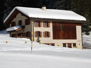 Eine zweistöckige Chalet mit schneebedecktem Dach und Holzläden, eingebettet in eine verschneite Landschaft.