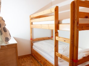 Un lit superposé avec une structure en bois et une literie blanche, dans une pièce dotée d’un plancher en bois et d’une petite table de chevet en bois.