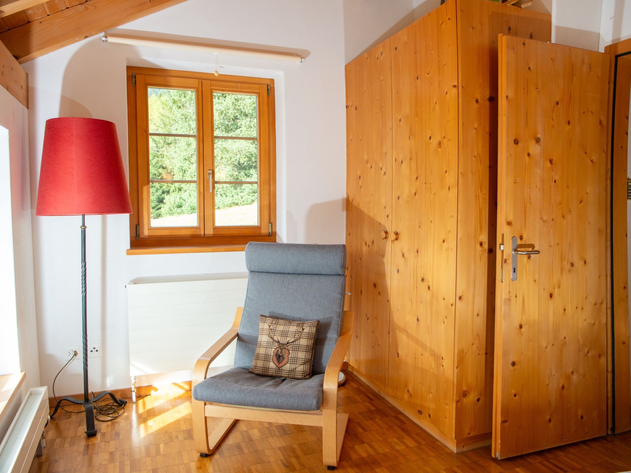 Une pièce avec une porte en bois, une fenêtre donnant sur un paysage verdoyant, un lampadaire rouge et une chaise bleue agrémentée d’un coussin à motifs.