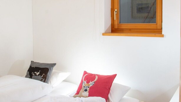 Um quarto com uma cama de solteiro, uma janela com moldura de madeira e uma luminária fixada na parede.