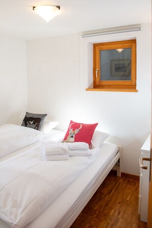 Un dormitorio con una cama individual, una ventana con marco de madera y una lámpara fijada a la pared.