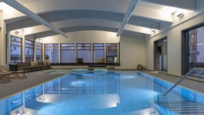 A large indoor swimming pool with clear water, a seating area with cushions, and a view of the outside through large windows. - 1620 Schweizerhof 402 by Interhome (Lenzerheide)