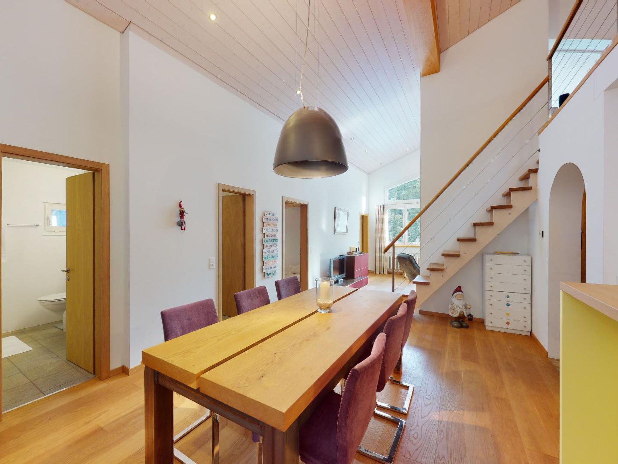 Een ruime eetruimte met een lange houten tafel, paarse stoelen en een moderne hanglamp.