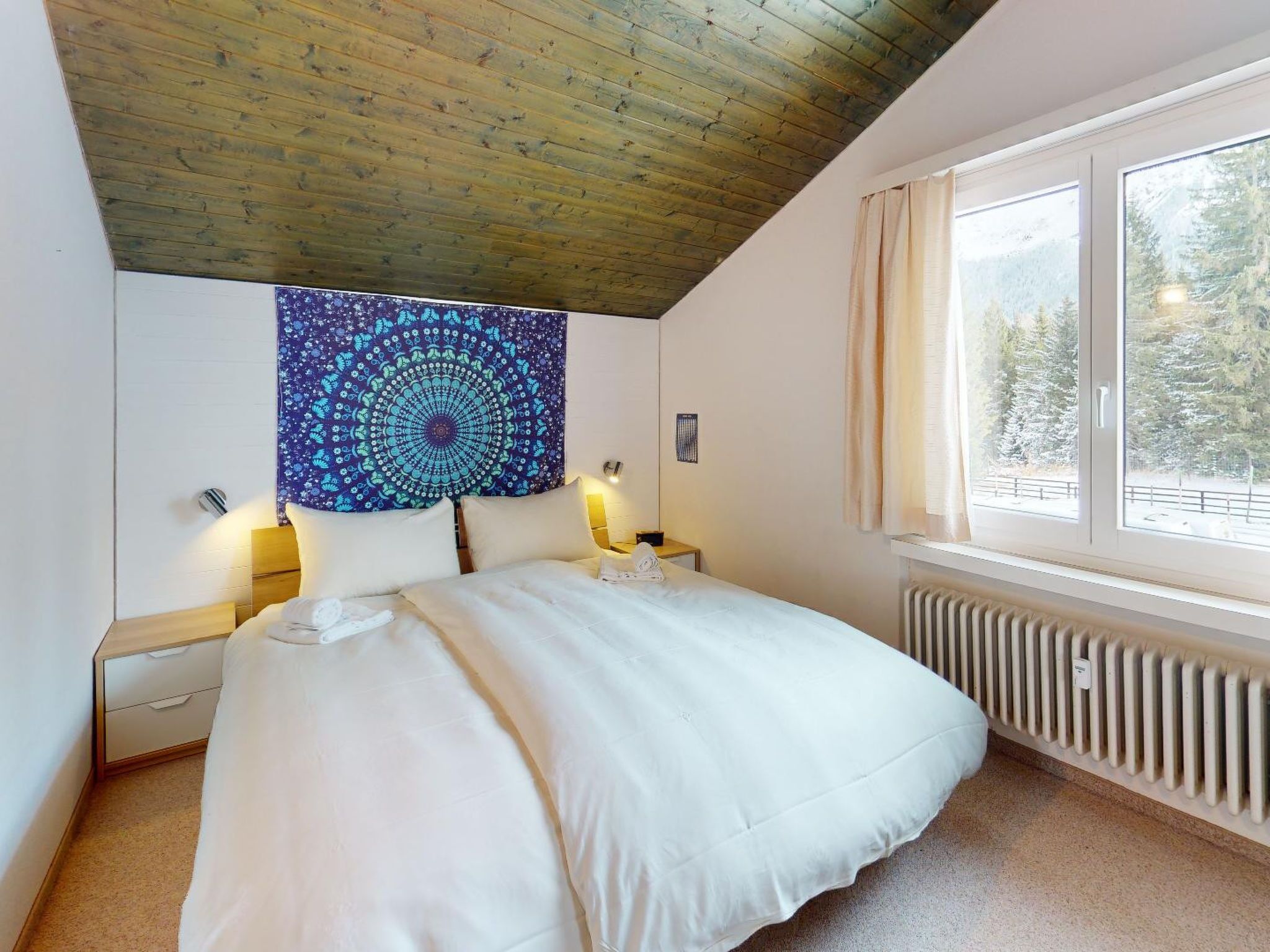 A bedroom with a large bed, a wooden ceiling, a tapestry, and a window with a view of trees.