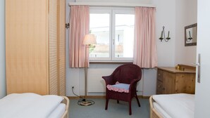 Una habitación de hotel con dos camas individuales, un armario de madera, una silla de mimbre, una mesita de noche con lámpara y una ventana con cortinas.
