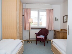 A hotel room with two single beds, a wooden wardrobe, a wicker chair, a bedside table with a lamp, and a window with curtains. - 1603 Sundrella by Interhome (Lenzerheide)