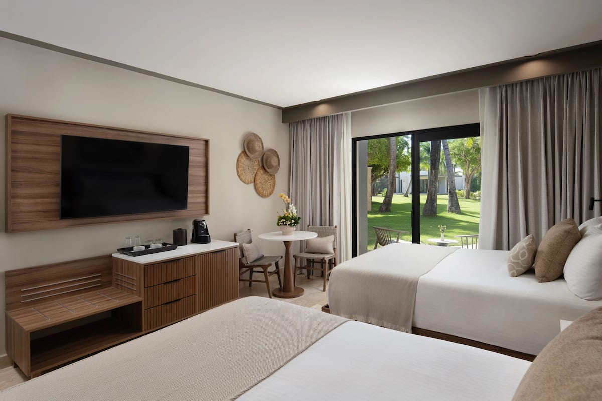 A modern hotel room with a large flat-screen TV, a desk with a coffee maker, two beds, a small round table with chairs, and a view of a green lawn through large windows.
