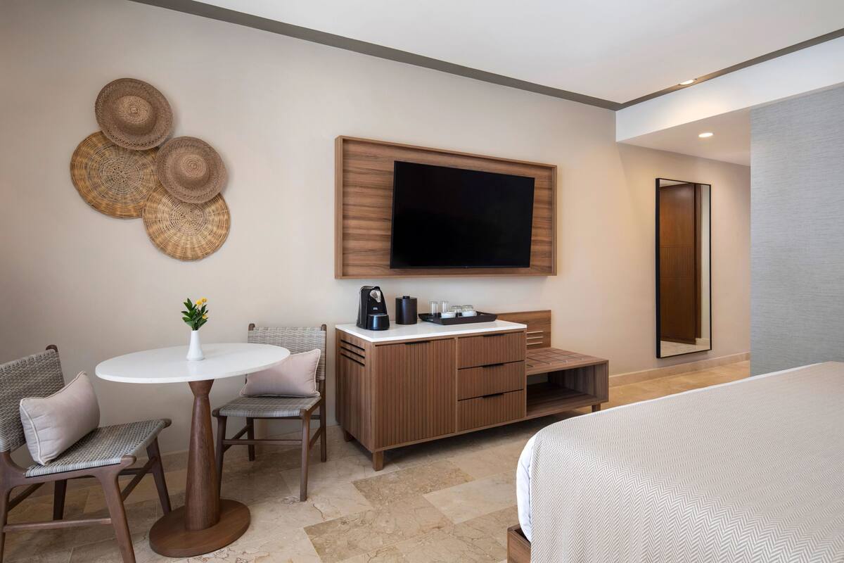 A hotel room with a bed, a TV mounted on the wall, a wooden cabinet, a small table with a vase, and two chairs.