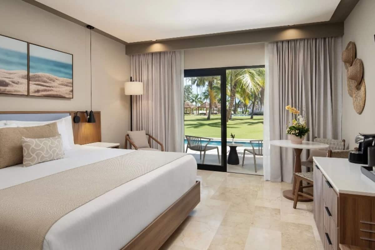 A hotel room with a bed, a desk, a chair, a small table with a plant, and a view of a pool and garden.
