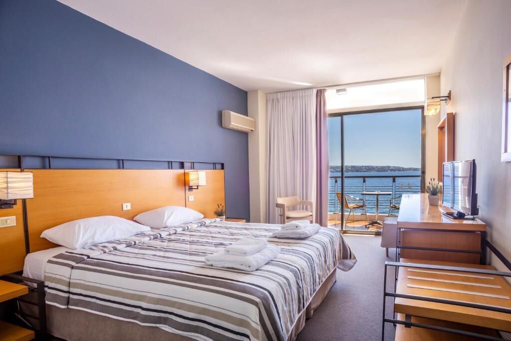 A hotel room with a bed, a desk, a chair, a TV, and a view of the sea.