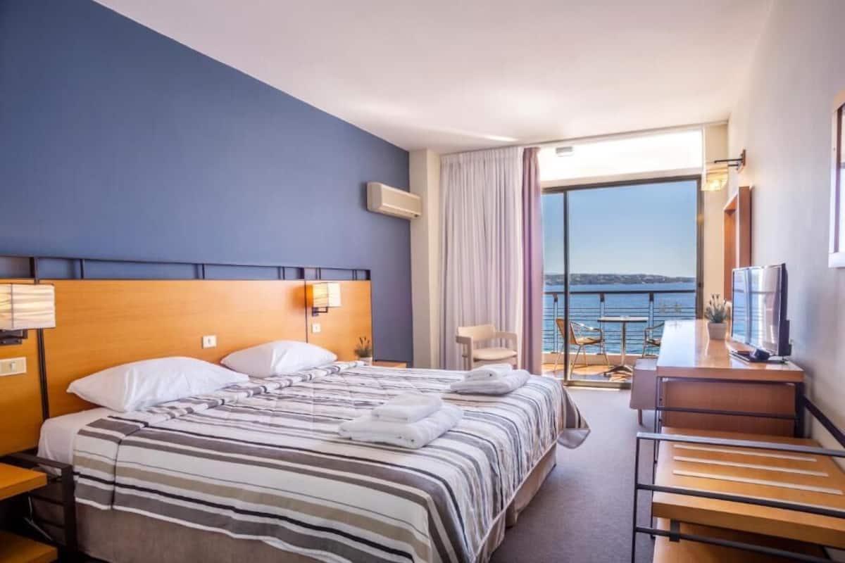 A hotel room with a bed, a desk, a chair, and a view of the sea.