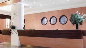 Un mostrador de recepción con diseño moderno, acabado en madera y separador de vidrio. Se observan tres ventanas redondas en la pared detrás del mostrador, y una planta en maceta se encuentra a la derecha.