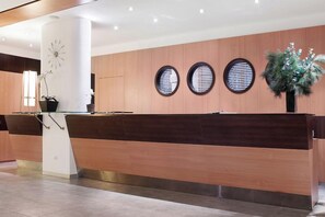 A reception desk with a modern design, featuring a wooden finish and a glass partition. There are three round windows on the wall behind the desk, and a potted plant is placed to the right. - Spetses (Spetses)
