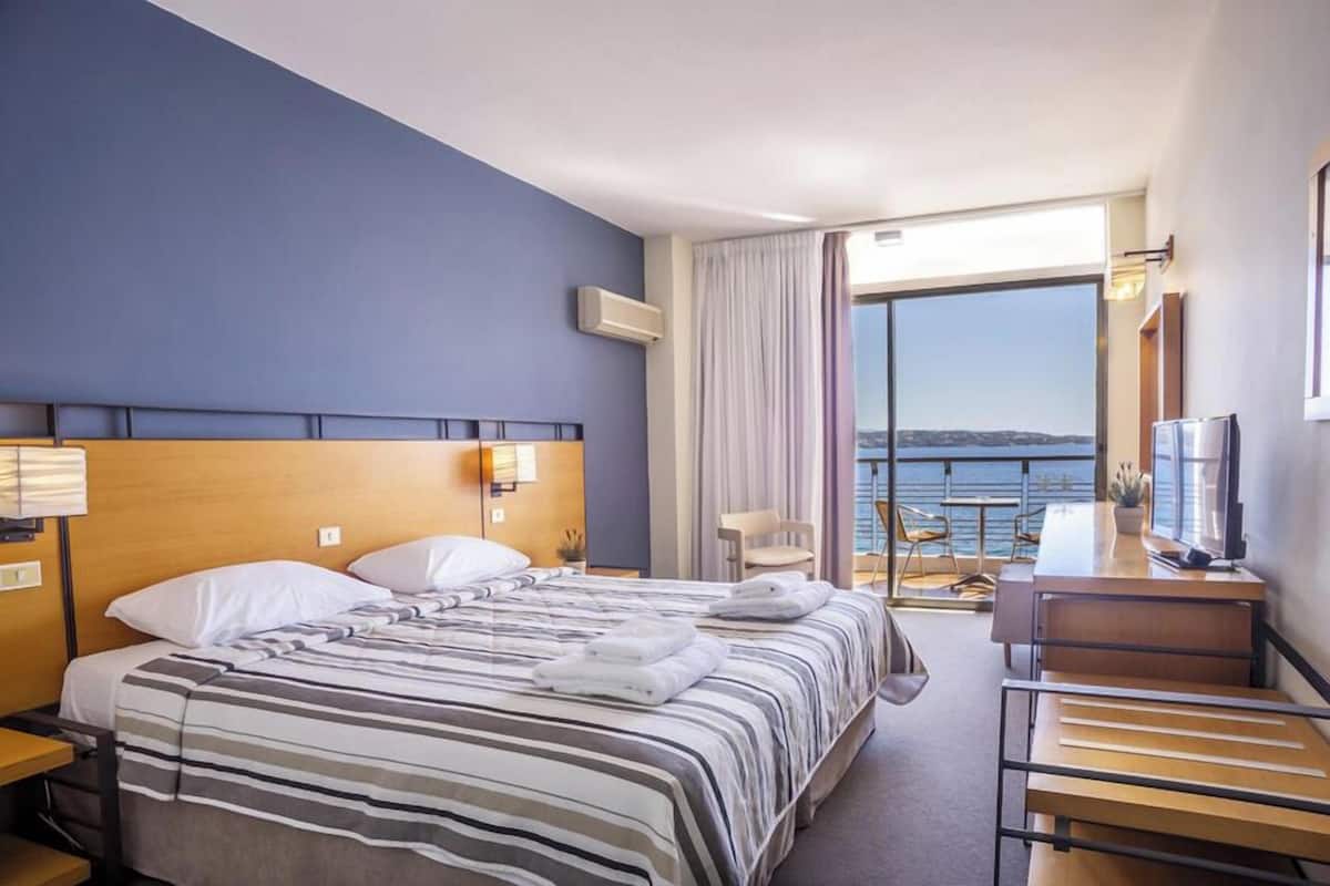 A hotel room with a bed, a desk, a chair, and a view of the sea.