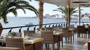 An outdoor dining area with wicker furniture, a view of the sea, and a cityscape in the background. - Spetses (Spetses)
