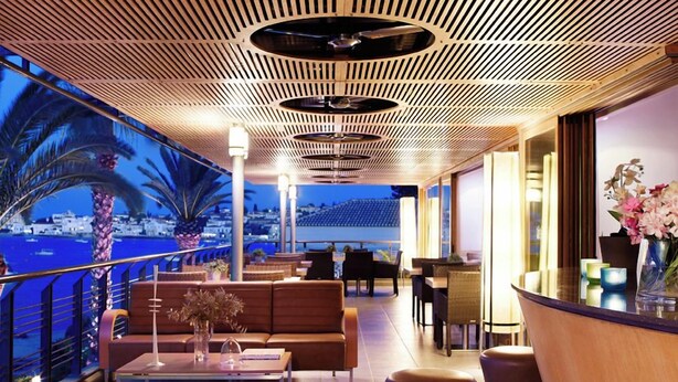 Un bar en la azotea con vistas al mar, distribución de mesas y un diseño de techo de madera.