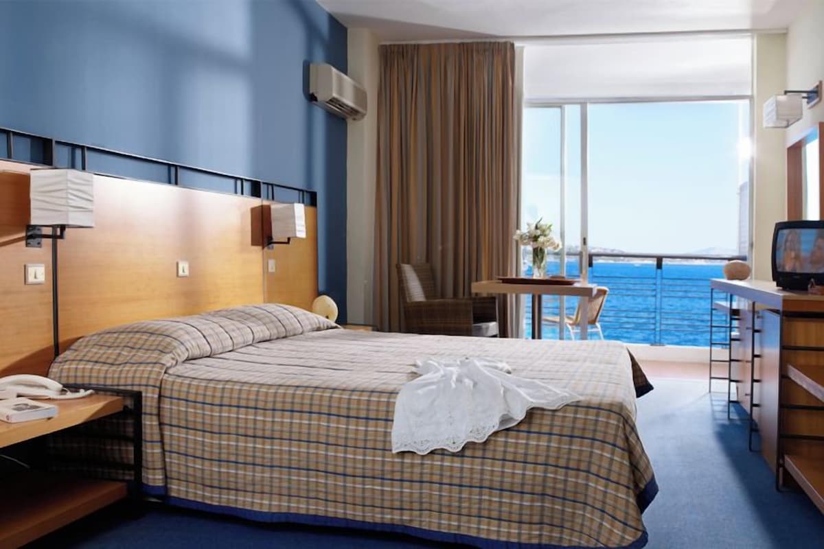 A hotel room with a large bed, a desk with a chair, a television, and a view of the ocean.