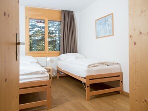 Une petite pièce propre et bien rangée, avec deux lits en bois, une fenêtre donnant sur des arbres, un tableau accroché au mur et une lampe de chevet.
