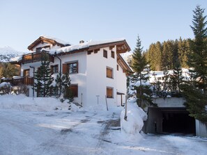 Un chalet à deux étages, avec un toit enneigé et des balcons en bois, entouré de neige et de pins.
