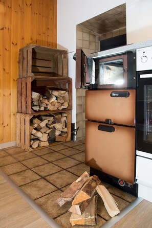 Una cocina con estufa de leña, leña apilada y un horno moderno.