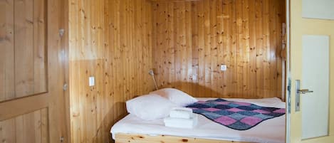 Un lit simple avec du linge de lit blanc et une couverture rose et bleue, dans une pièce en bois avec une porte sur le côté droit.