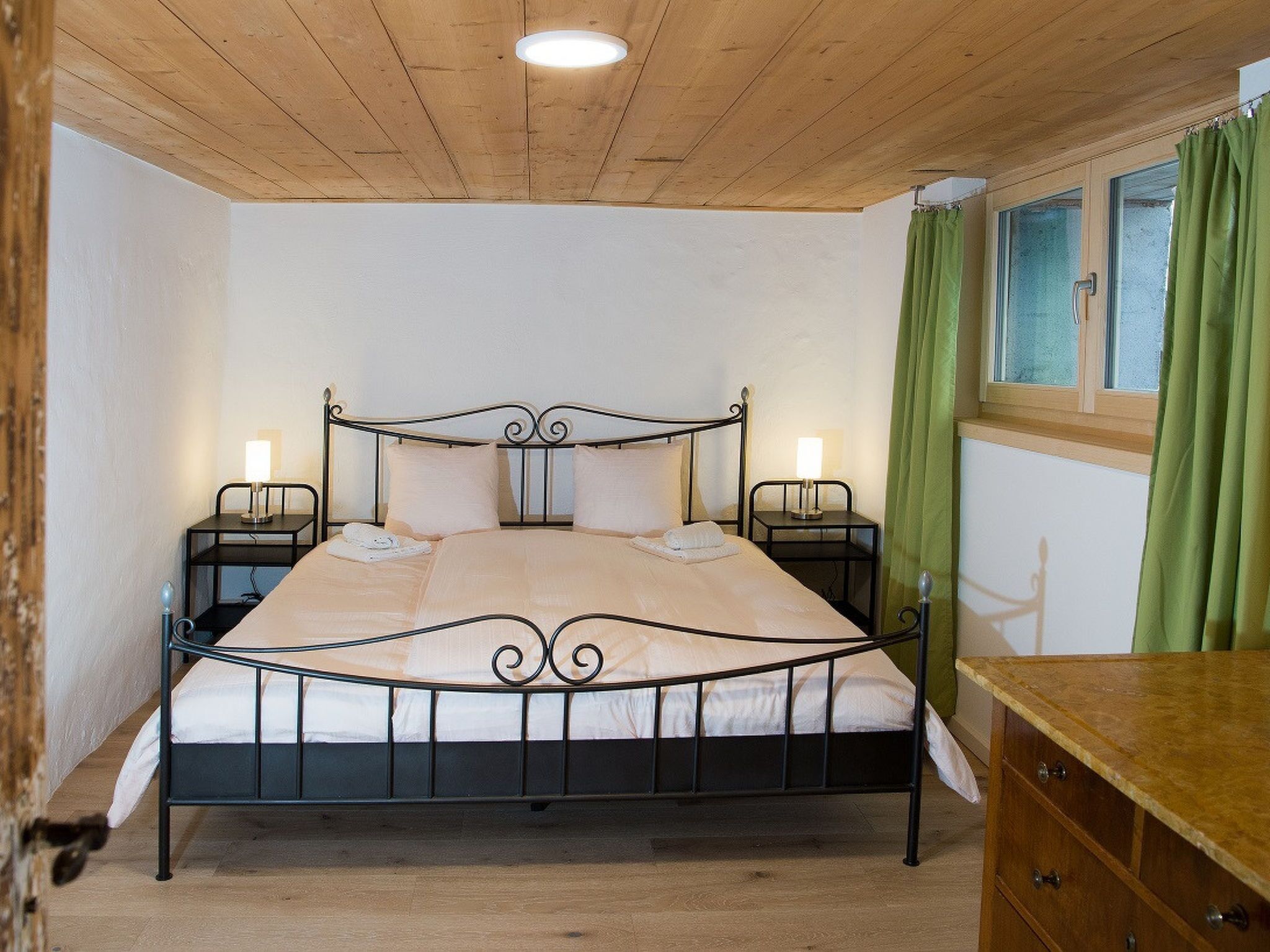 A single bed with a metal frame, two bedside tables with lamps, a wooden ceiling, and a window with green curtains.